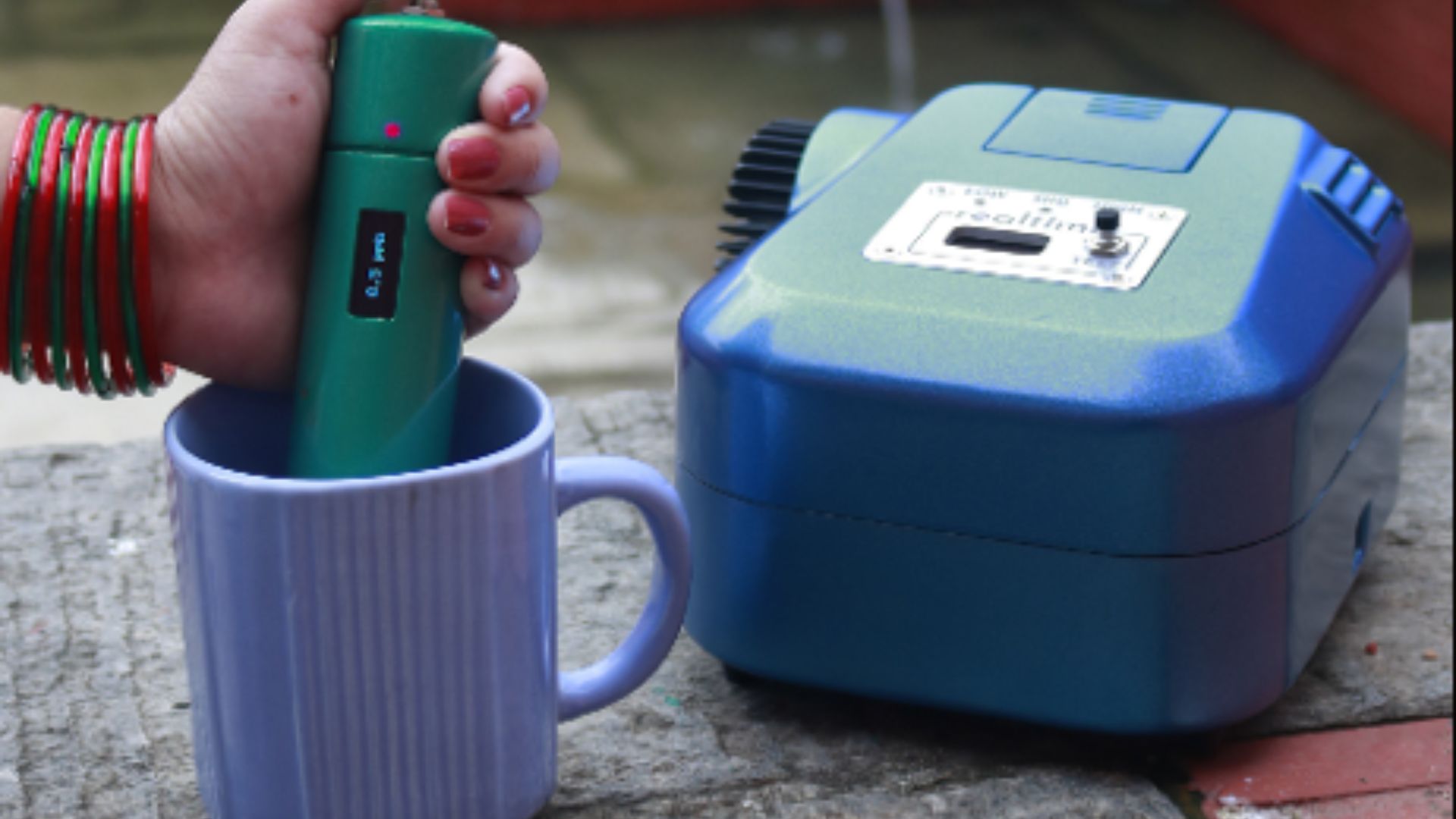 Dispositivo portátil de detección de contaminantes en el agua, de color verde, siendo sumergido en una taza mientras está conectado a un equipo de análisis más grande, de color azul. La mano de una persona sostiene el dispositivo verde, que muestra una pantalla digital pequeña.