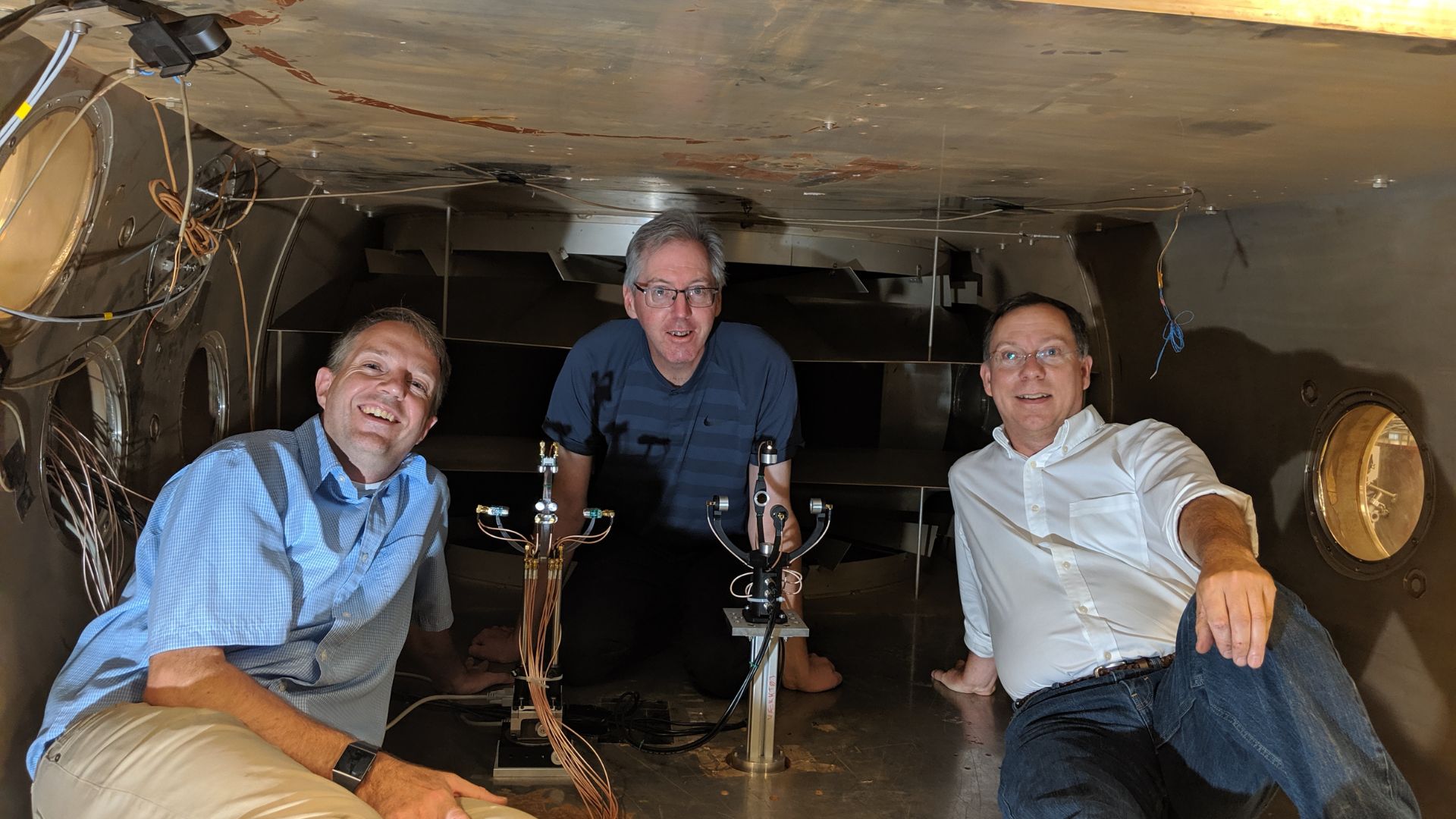 Tres hombres sentados en el interior de una cámara metálica grande, posando junto a un equipo científico. El dispositivo, situado entre ellos, parece ser parte de un sistema experimental, posiblemente relacionado con la medición de viento. Los tres están vestidos casualmente y parecen estar trabajando en un entorno de investigación.