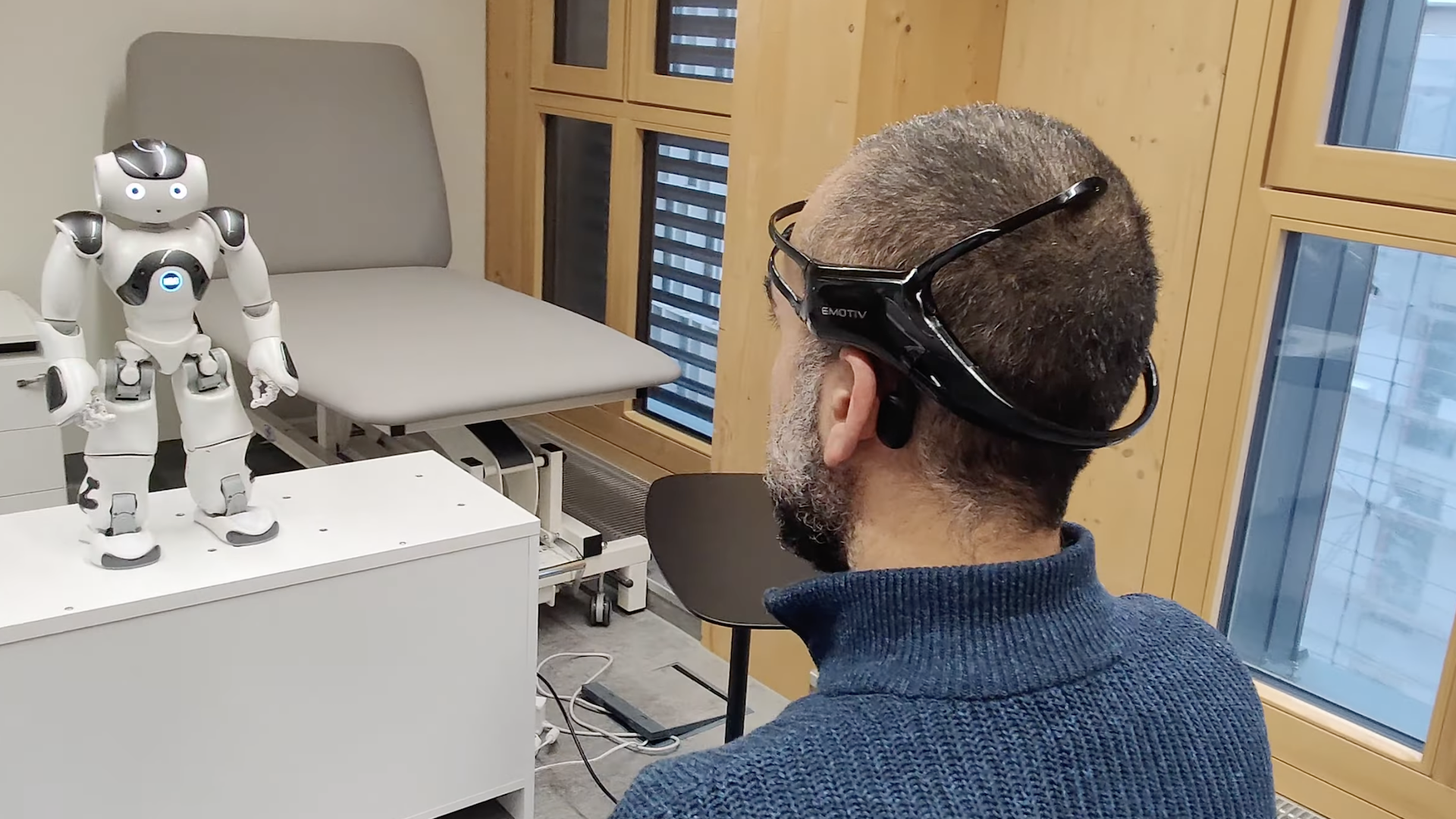 Un hombre con un casco de interfaz cerebro-computadora (BCI) está sentado frente a un pequeño robot humanoide en una sala de terapia. El robot, que está sobre una mesa blanca, parece estar interactuando con el hombre. La escena sugiere un entorno de rehabilitación, posiblemente relacionado con un estudio en el que se utilizan robots asistenciales para ayudar a pacientes mediante la detección de la actividad neural. Al fondo se observa una camilla médica y una ventana.