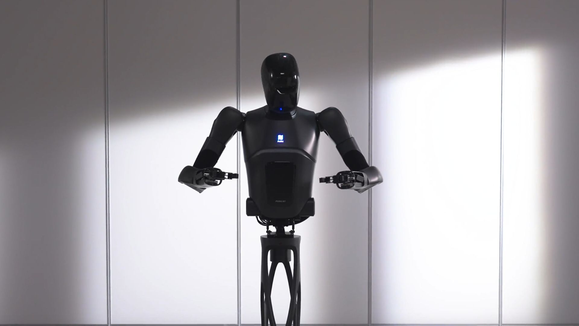 Un robot semi-humanoide de color negro desarrollado por Pudu Robotics. El robot tiene un torso con una estructura robusta y brazos robóticos con manos capaces de agarrar objetos. Su diseño incluye una cabeza sin rasgos faciales visibles, dándole una apariencia futurista y minimalista. Está situado en una sala con paredes blancas, iluminado parcialmente desde un lado, lo que resalta su figura y detalles tecnológicos.