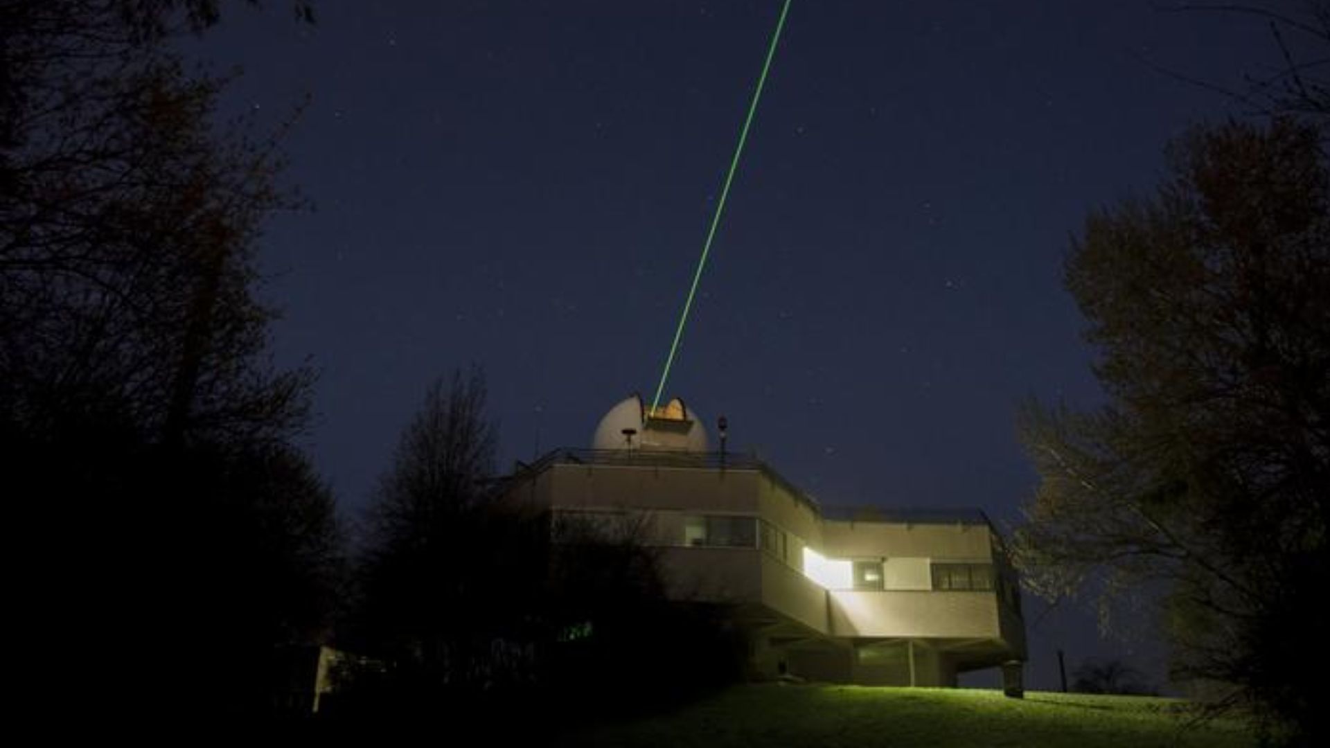 Observatorio en funcionamiento durante la noche, con un láser verde apuntando hacia el cielo desde la cúpula del edificio. Alrededor, se ven árboles y el entorno oscuro, con una luz tenue en el observatorio. La imagen representa la técnica de Satellite Laser Ranging (SLR), utilizada para rastrear satélites en órbita mediante la reflexión de láseres.