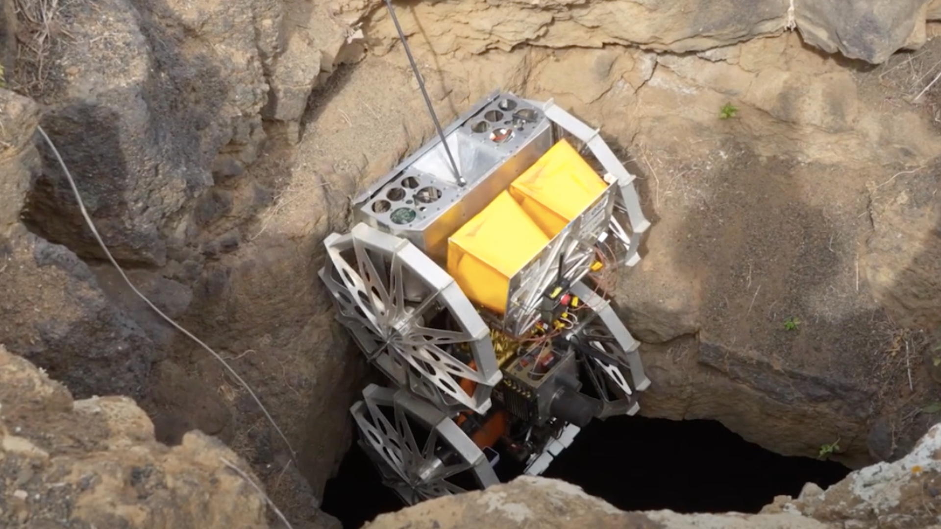 Robot explorador descendiendo por una abertura natural (skylight) hacia un tubo de lava durante una prueba en terrenos volcánicos. La máquina está equipada con ruedas robustas y un sistema de cabrestante que asegura un descenso controlado, diseñado para simular condiciones de exploración lunar.